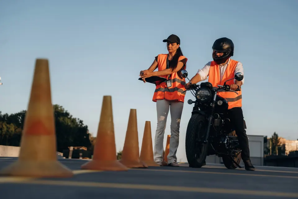 Élève motard casqué en gilet orange sur une piste de plots, avec sa monitrice